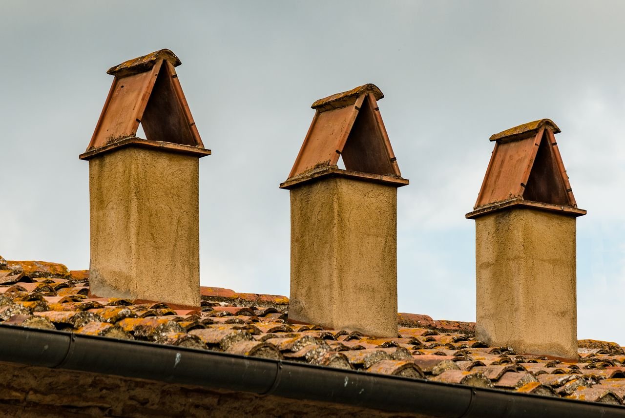 why-choose-us-03 Image of three chimneys with triangular covers on a tiled roof under an overcast sky.
