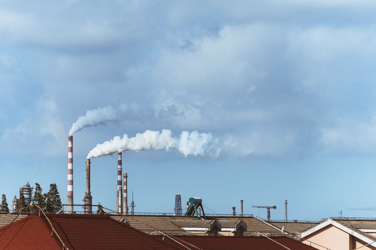 our-mission Factory chimneys emitting smoke over rooftops on a clear day, illustrating urban pollution.